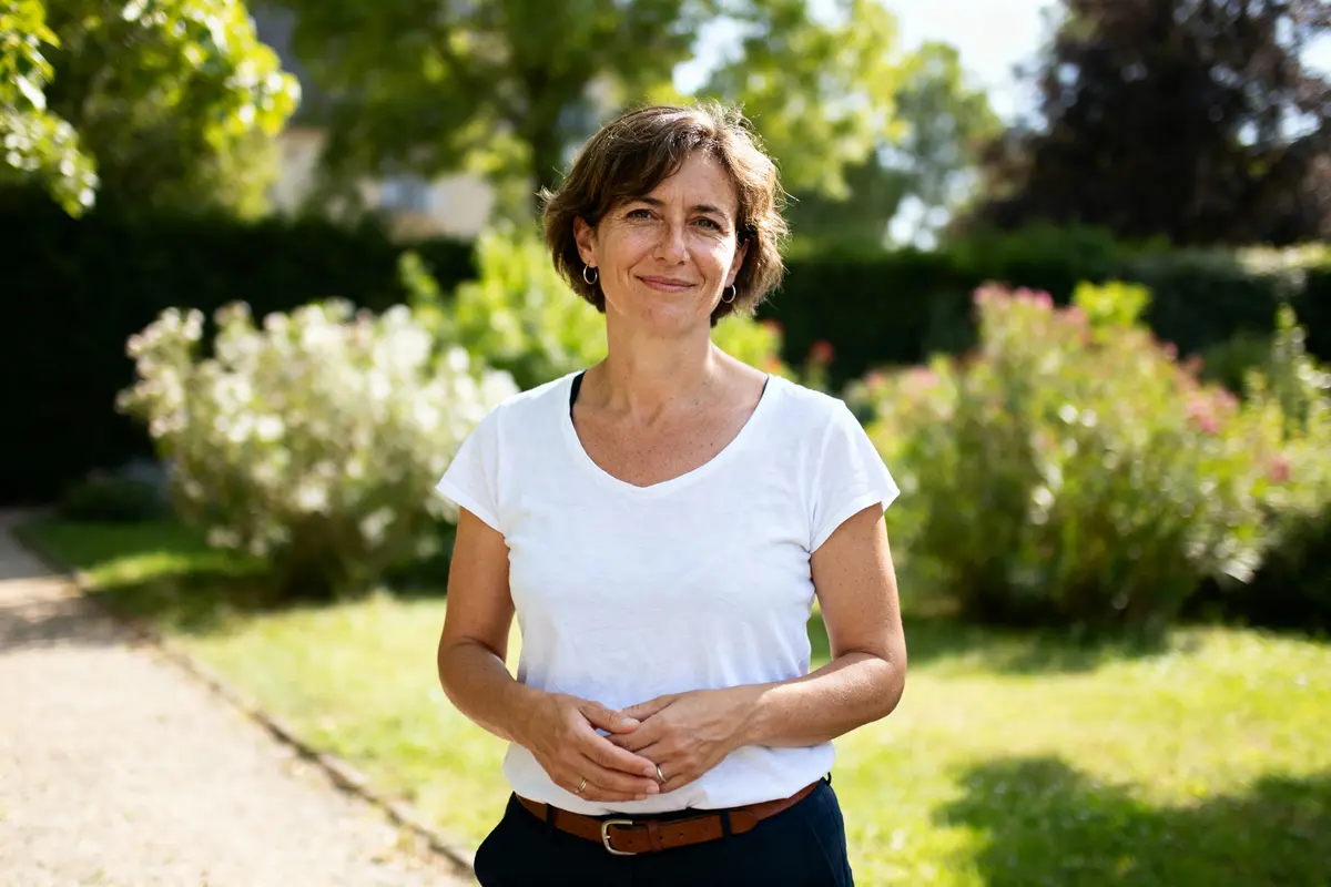 Femme souriante en t-shirt blanc, debout dans un jardin verdoyant et ensoleillé.