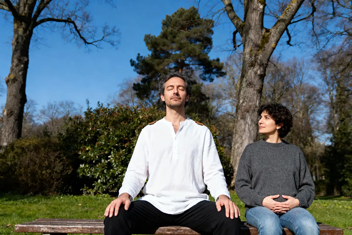 Deux personnes assises sur un banc, l'homme médite, la femme regarde, arbres et ciel bleu en arrière-plan.