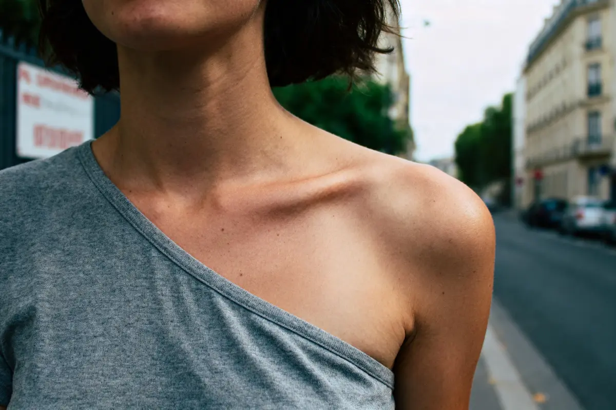 Femme en haut gris à une épaule, dans une rue urbaine avec immeubles flous en arrière-plan.
