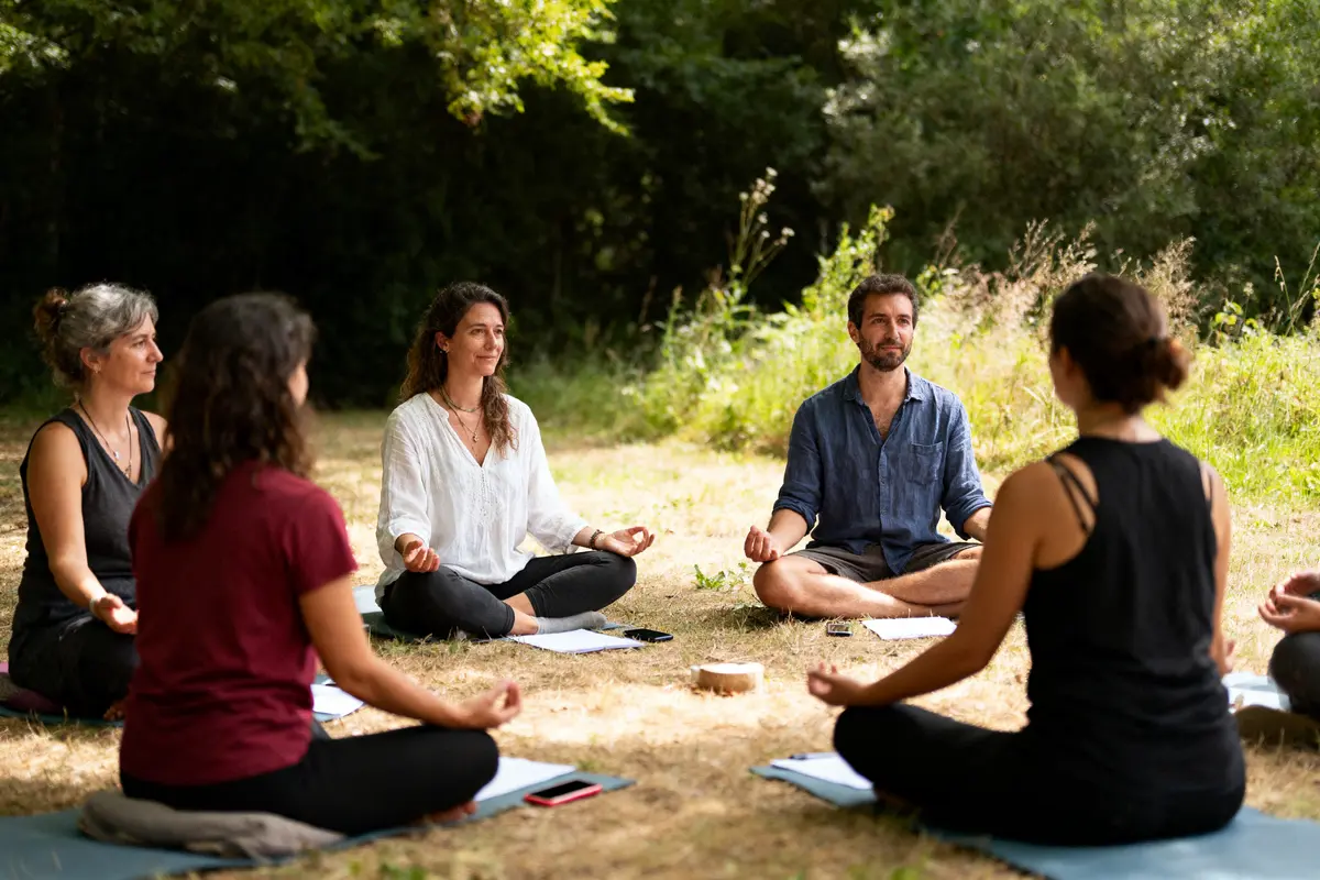 Personnes méditant en cercle en plein air, assises sur des tapis, entourées de végétation. Ambiance paisible et naturelle.