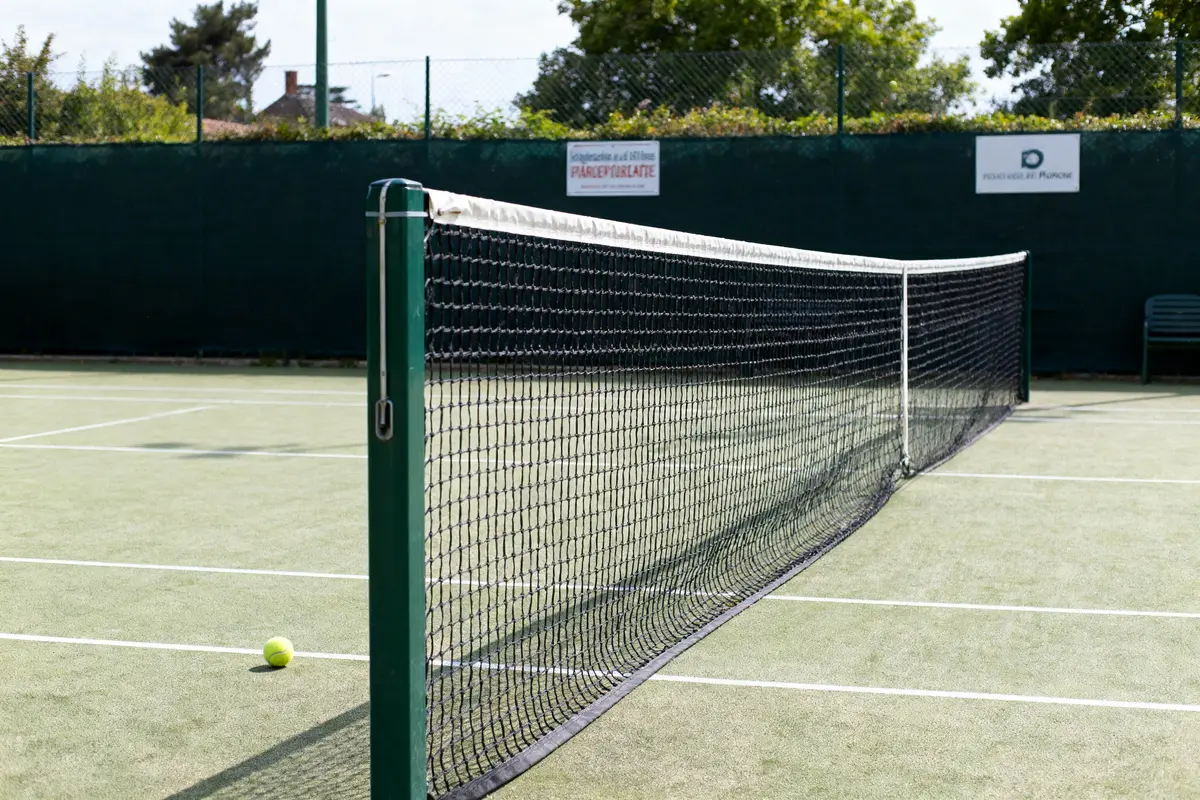 Terrain de tennis avec filet et balle jaune sur surface engazonnée, clôturé par un grillage.