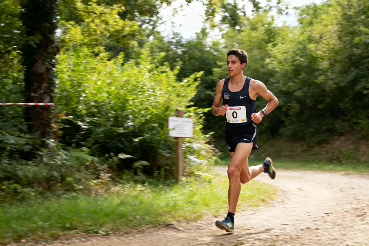 Coureur en tenue sportive noire, numéro 0, sur un sentier forestier. Arbres verts en arrière-plan.