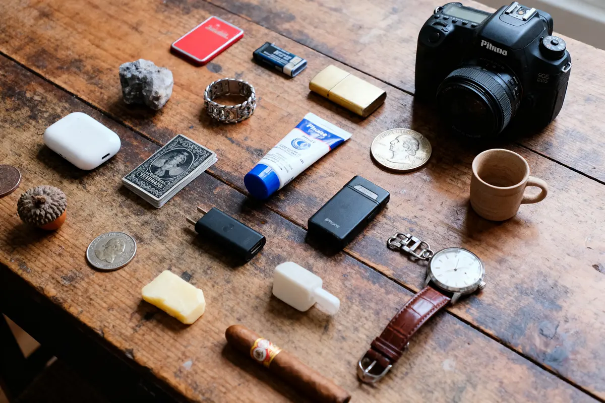 Objets variés sur table en bois incluant appareil photo, montre, tube, pièce, carnet, bague, USB, gobelet.