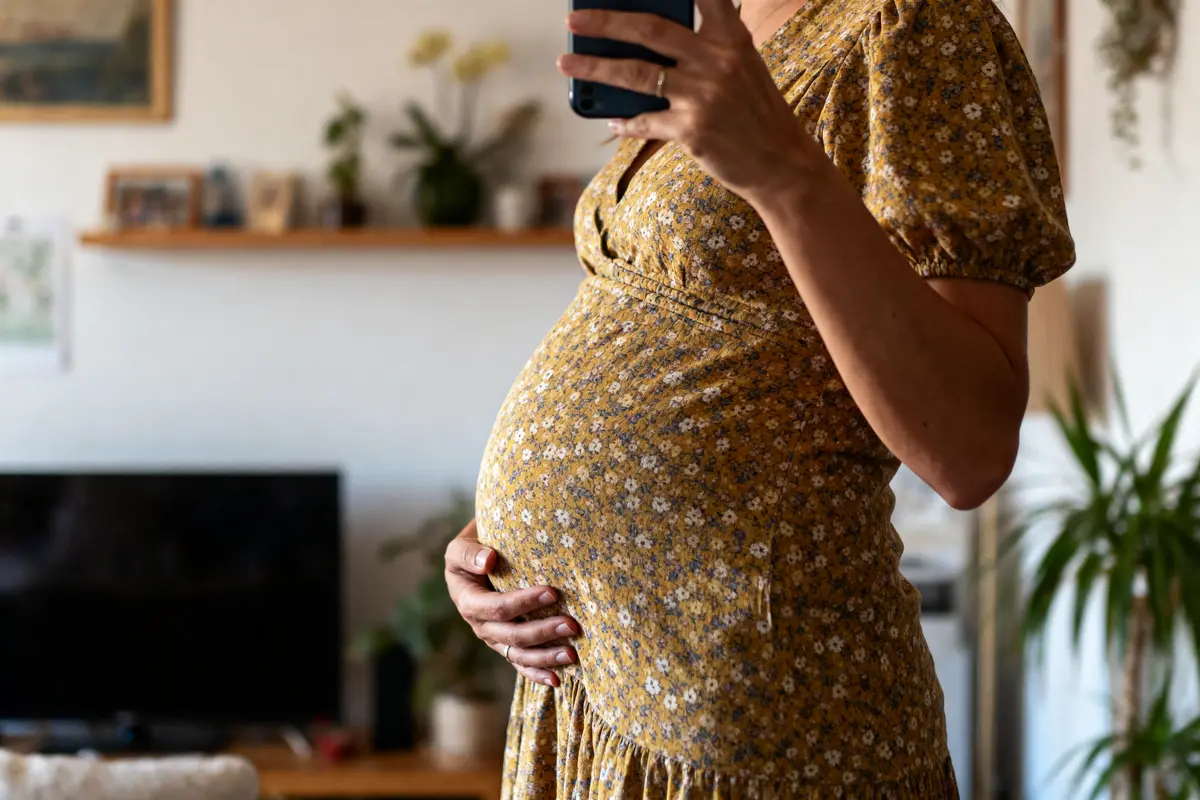 Femme enceinte en robe à fleurs, tenant son ventre et un téléphone, intérieur avec plantes.