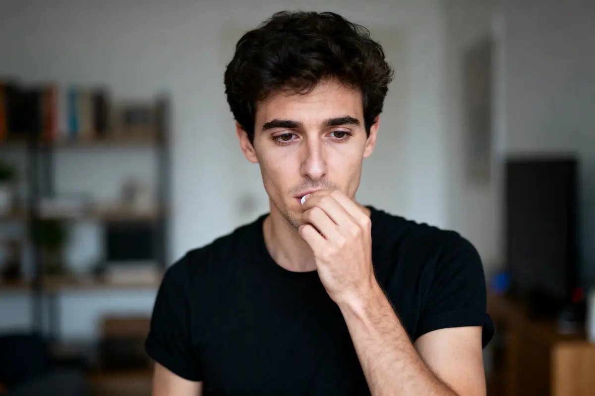 Homme pensif en noir, main sur la bouche, intérieur flou en arrière-plan.