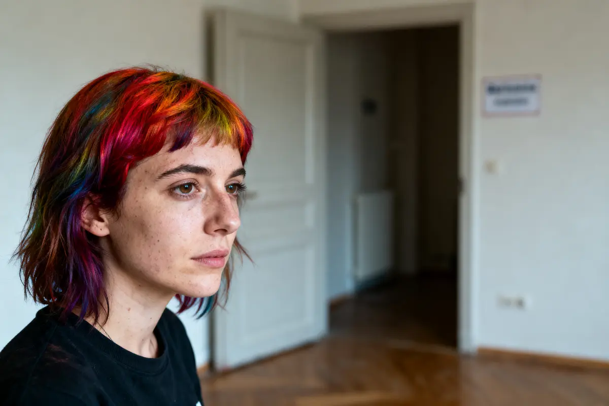 Personne aux cheveux multicolores regarde à gauche dans une pièce lumineuse avec parquet et porte entrouverte.