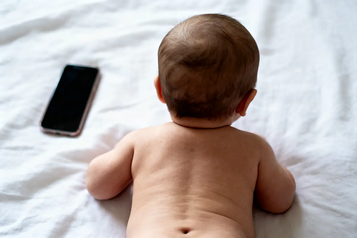 Bébé allongé sur le ventre, regardant un smartphone sur un lit blanc.