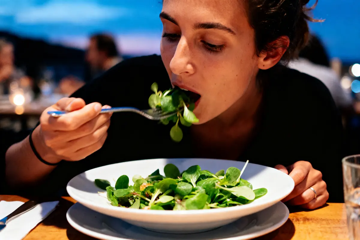 Personne mangeant une salade verte avec une fourchette, assise à une table en soirée à l'extérieur.