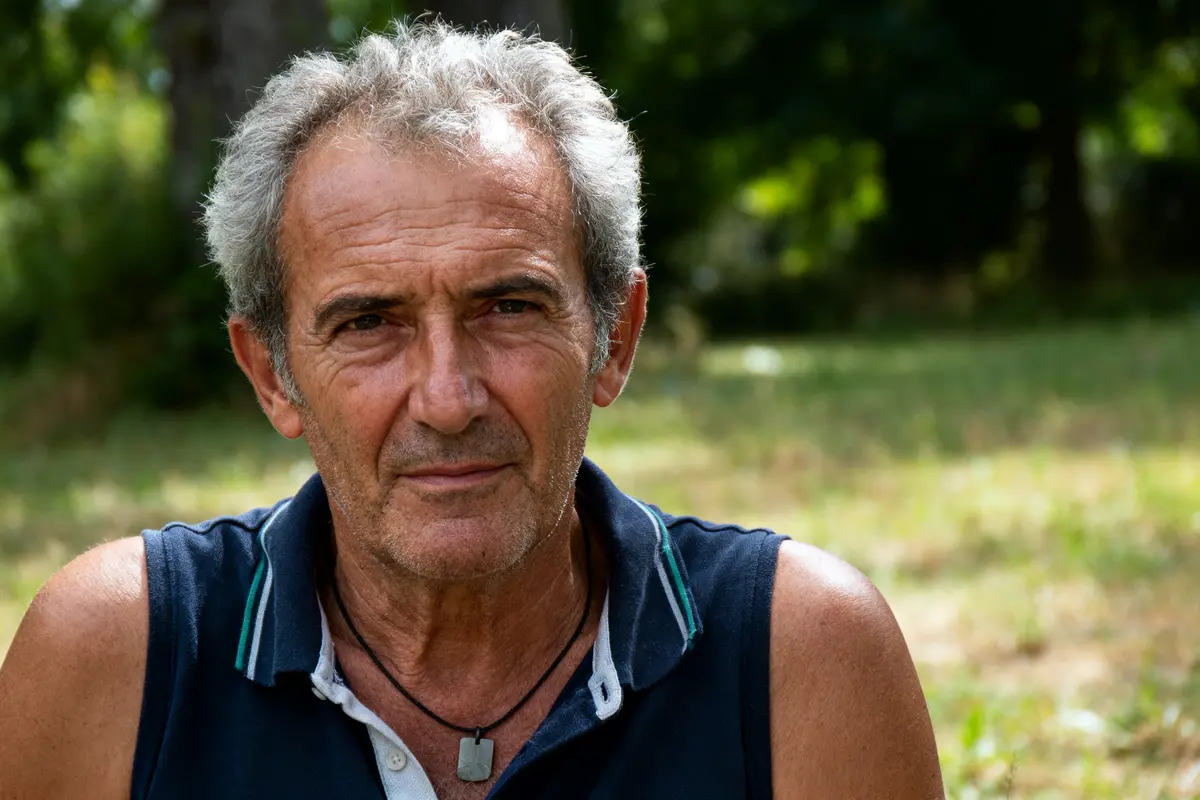 Homme âgé avec cheveux gris, expression sérieuse, fond flou verdoyant, portant débardeur bleu et collier noir.