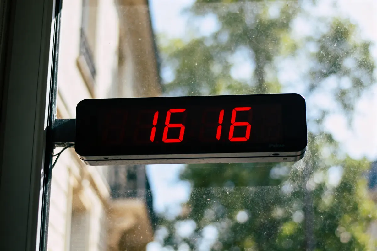 Horloge numérique avec affichage LED rouge indiquant 16h16, montée sur une fenêtre. Arrière-plan flou extérieur.