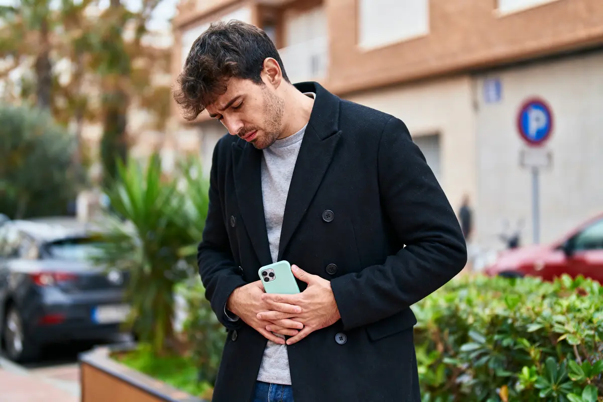 Un homme avec un manteau noir tient son ventre, l'air préoccupé, avec un téléphone en main.