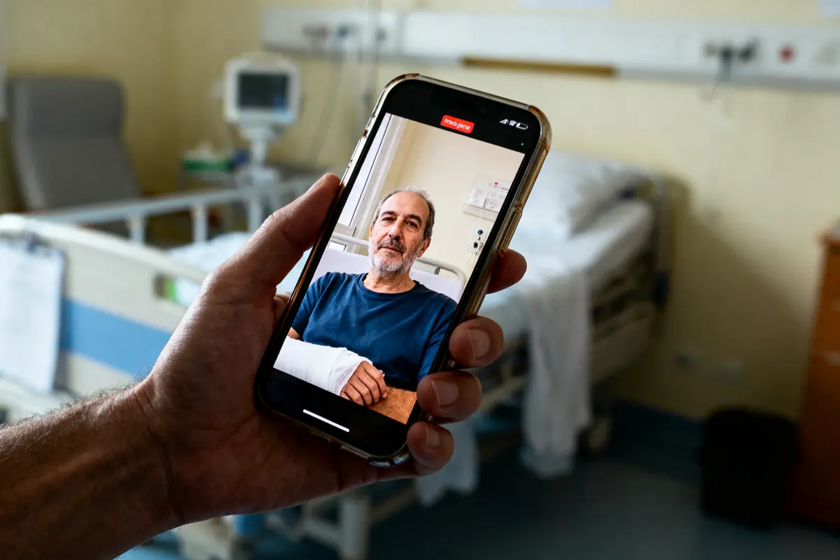 Photo d'un homme, bras bandé, sur téléphone devant un lit d'hôpital vide.