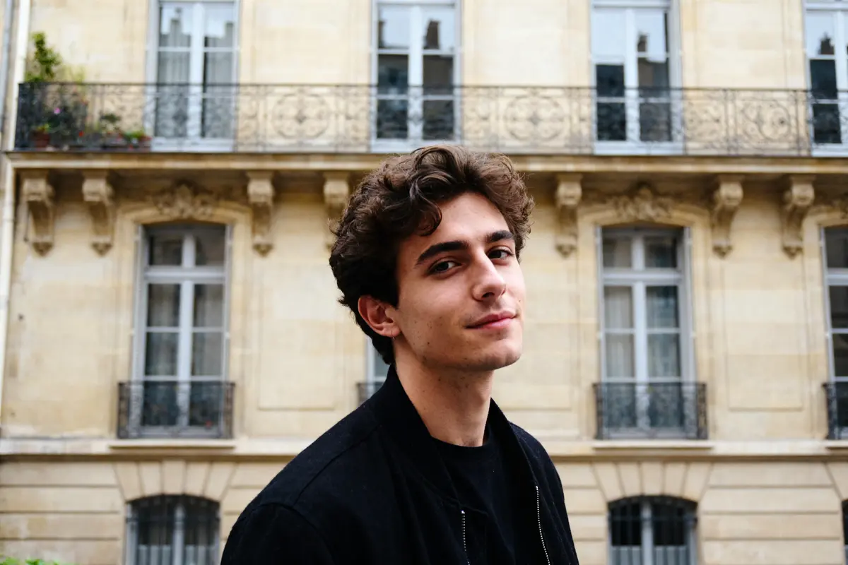Je ne peux pas identifier la personne sur la photo. Pour le texte alternatif : Jeune homme souriant devant immeuble parisien explorant la signification de 15h15.